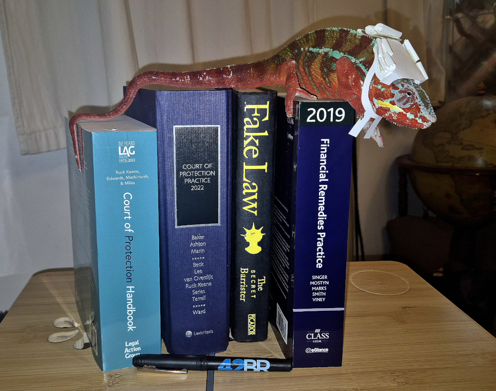 A colourful lizard in a tiny paper barrister wig and bands lies across a pile of law books